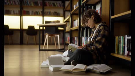 Una-Chica-Morena-Concentrada,-Con-Cabello-Rizado-Y-Gafas,-Lee-Un-Libro-En-La-Biblioteca-Entre-Los-Estantes-Y-Los-Libros-En-El-Suelo.