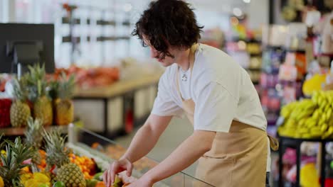 A-confident-guy-a-brunette-store-worker-with-curly-hair-in-a-white-T-shirt-and-a-yellow-apron-lays-out-goods-on-the-counter-in-a-supermarket