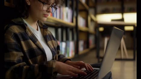 Close-up-a-girl-student-with-glasses-with-curly-hair-in-a-checkered-shirt-makes-notes-in-a-gray-laptop-looking-at-the-shelves-with-books-at-the-university