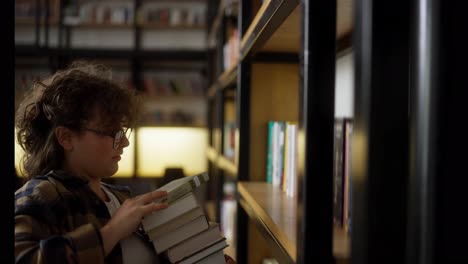 A-girl-student-with-curly-hair-with-glasses-in-a-checkered-shirt-puts-a-stack-of-books-on-a-shelf-in-the-library