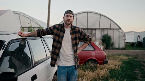 Portrait-of-a-serious-farmer-guy-with-a-beard-in-a-checkered-shirt-posing-against-of-greenhouses-on-the-farm