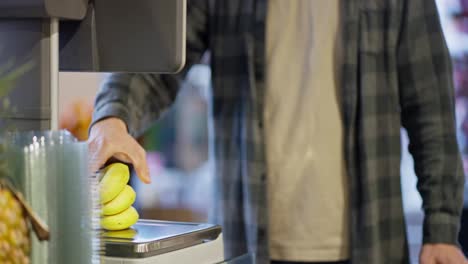 Close-up-Supermarket-visitor-weighs-bananas-using-scales-in-supermarket.-A-guy-in-a-plaid-shirt-uses-a-scale-to-weigh-bananas-while-shopping-in-a-supermarket