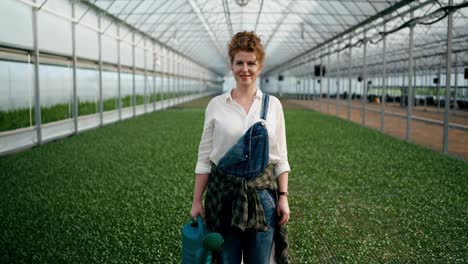 Portrait-of-a-happy-girl-with-curly-hair-in-a-white-farmer-shirt-with-a-watering-can-in-her-hands-on-a-farm-in-a-plant-greenhouse