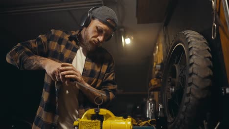 Un-Mecánico-Con-Barba,-Gorra-Y-Auriculares-Trabaja-Con-Una-Lima-En-Un-Banco-De-Trabajo-Y-Escucha-Música-Mientras-Trabaja-En-Un-Taller-De-Estudio.