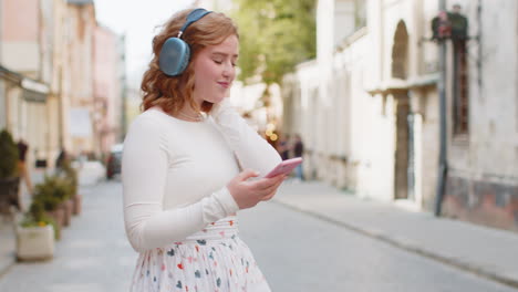 Happy-young-woman-in-wireless-headphones-choosing,-listening-music-in-smartphone-dancing-outdoors