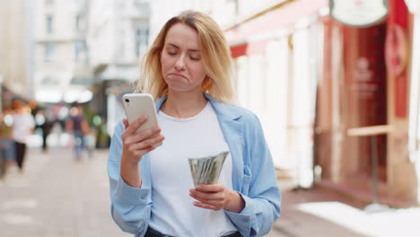 Mujer-Rubia-Feliz-Contando-Dinero-En-Efectivo-En-Dólares,-Usa-La-Aplicación-De-Calculadora-De-Teléfono-Inteligente-En-La-Calle-De-La-Ciudad