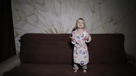 Little-cute-girl-with-blond-hair-jumps-on-sofa.-Bathrobe-clothes.-Happy