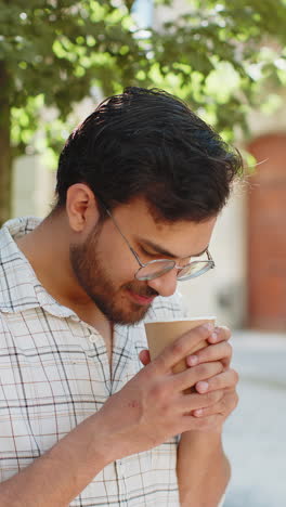 Happy-Indian-young-man-enjoying-morning-coffee-hot-drink-and-smiling-standing-on-city-street