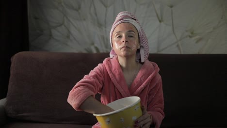 Woman-watching-a-late-night-movie-at-TV,-eating-popcorn.-Bathrobe,-facial-mask