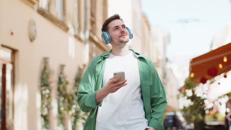 Caucasian-man-tourist-in-wireless-headphones-listening-music-in-smartphone-walking-sunny-city-street