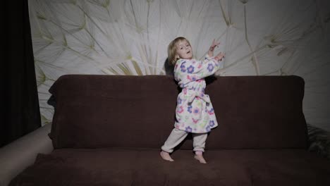 Little-cute-girl-with-blond-hair-jumps-on-sofa.-Bathrobe-clothes.-Happy