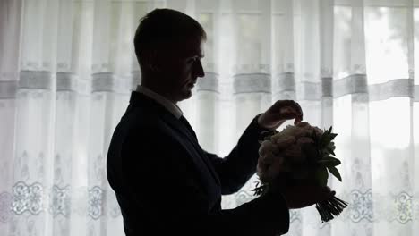 Handsome-groom-fixes-his-wedding-bouquet-near-window.-Silhouette