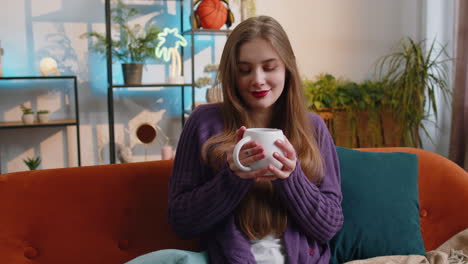 Pretty-smiling-young-woman-drinking-a-cup-of-coffee-or-herbal-tea-sitting-at-home-couch,-break-time