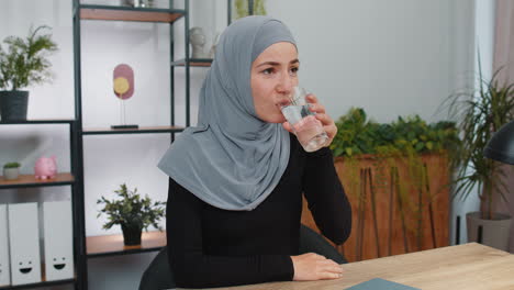 Thirsty-muslim-woman-sitting-at-home-holding-glass-of-natural-aqua-make-sips-drinking-still-water