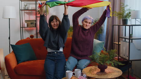 Two-lesbian-women-family-couple-or-girl-friends-holding-LGBT-people-gay-pride-flag-in-hands-at-home