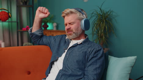 Happy-mature-man-in-wireless-headphones-relaxing-sitting-on-sofa-at-home-listening-favorite-music