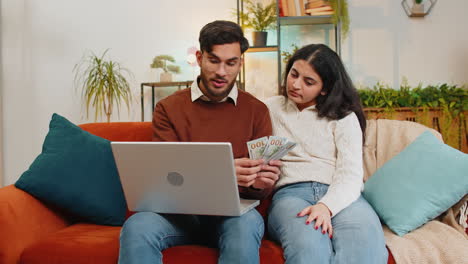 Happy-Indian-couple-counting-money-and-booking-tickets-for-holiday-vacation-sitting-on-sofa-at-home