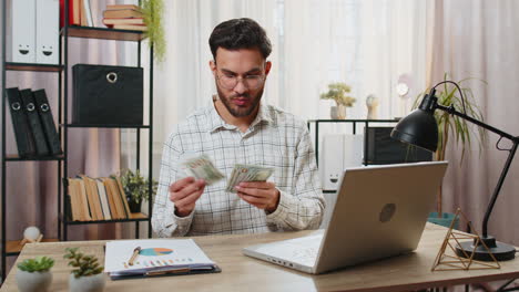 Indian-business-man-counting-money-cash-calculates-dividends-and-using-laptop-in-home-office