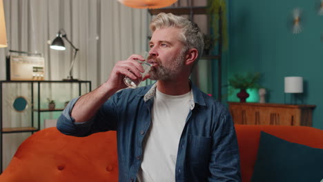 Thirsty-middle-aged-man-sitting-at-home-holding-glass-of-natural-aqua-make-sips-drinking-still-water