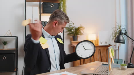 Frustrated-young-businessman-in-formal-suit-with-clock-and-sticky-notes-sitting-at-home-office-desk