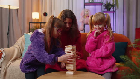 Three-siblings-children-girls-playing-with-blocks-board-game,-build-tower-from-wooden-bricks-at-home