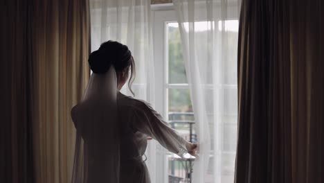 Bride-in-white-boudoir-dress-going-to-balcony-and-opens-curtains-and-door-in-hotel-room-apartment