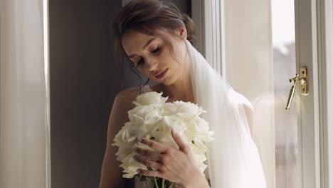 Bride-in-white-dress-staying-near-window-with-flowers-bouquet,-wedding-morning-preparations-at-home