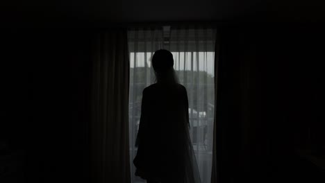 Lovely-bride-in-white-boudoir-dress-goes-to-balcony-and-opens-curtains-in-hotel-room-or-apartment