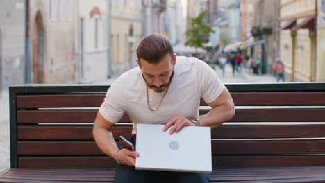 Junger-Freiberufler-Beginnt-Am-Laptop-Zu-Arbeiten,-Sendet-Nachrichten,-Tätigt-Online-Einkäufe-Auf-Der-Stadtbank