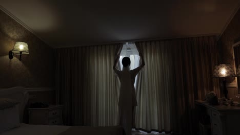 Bride-in-white-boudoir-dress-stay-near-window-and-opening-curtains,-woman-in-night-gown-and-veil