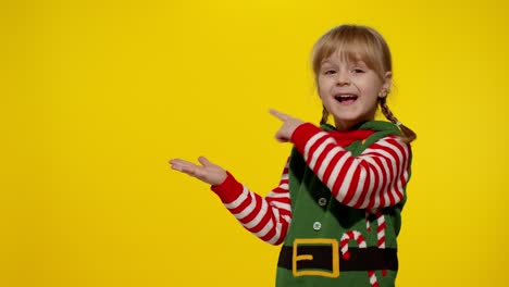 Kid-girl-in-Christmas-elf-Santa-helper-costume-points-fingers-at-blank-space-shows-advertising-area