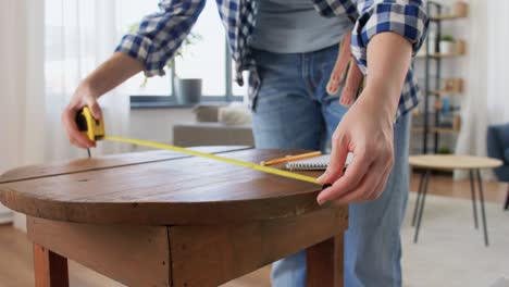 Woman-with-Ruler-Measuring-Table-for-Renovation.repair,-diy-and-home-improvement-concept--woman-with-ruler-measuring-old-round-wooden-table-for-renovation-and-writing-to-notebook