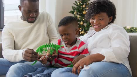 Afrikanische-Familie-Mit-Spielzeug-An-Weihnachten-Zu-Hause.Familie,-Winterferien-Und-Menschenkonzept.-Glückliche-Afroamerikanische-Mutter,-Vater-Und-Kleiner-Sohn-Mit-Dinosaurierspielzeug-Zu-Hause-An-Weihnachten