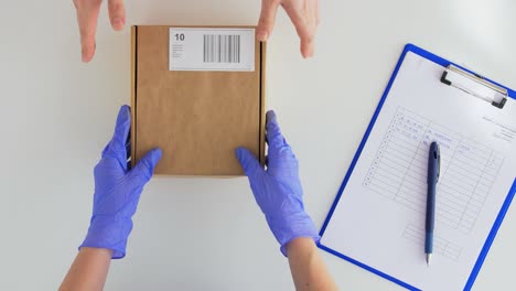 Hands-in-Gloves-with-Parcel-Box-and-Clipboard.health-protection,-safety-and-pandemic-concept--hands-of-delivery-woman-in-gloves-with-clipboard-giving-parcel-box-to-customer