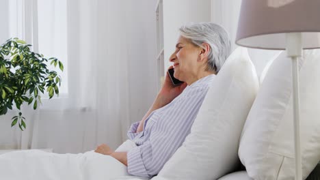 Senior-Woman-Calling-on-Smartphone-in-Bed-at-Home.old-age,-technology-and-people-concept--happy-smiling-senior-woman-in-pajamas-calling-on-smartphone-sitting-in-bed-at-home-bedroom