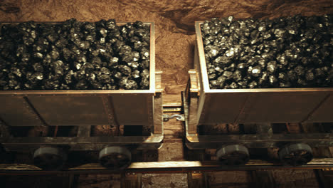 Old-mine-wagons-full-of-coal-moving-slowly-through-the-narrow-passage.-Seamless-looping-top-view-of-an-endless-row-of-coal-trolleys-in-a-dark-underground-tunnel.-Bright-light-at-the-end-of-tunnel.