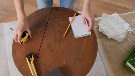 Woman-with-Ruler-Measuring-Table-for-Renovation.repair,-diy-and-home-improvement-concept--woman-with-ruler-measuring-old-round-wooden-table-for-renovation-and-writing-to-notebook