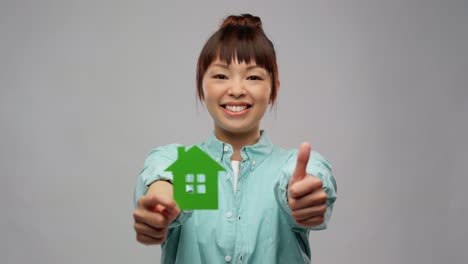 Asian-Woman-with-Green-House-Showing-Thumbs-Up.eco-living,-environment-and-sustainability-concept--portrait-of-happy-smiling-young-asian-woman-in-turquoise-shirt-holding-green-house-and-showing-thumbs-up-over-grey-background