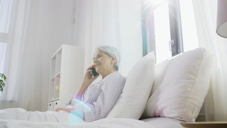 Senior-Woman-Calling-on-Smartphone-in-Bed-at-Home.old-age,-technology-and-people-concept--happy-smiling-senior-woman-in-pajamas-calling-on-smartphone-sitting-in-bed-at-home-bedroom