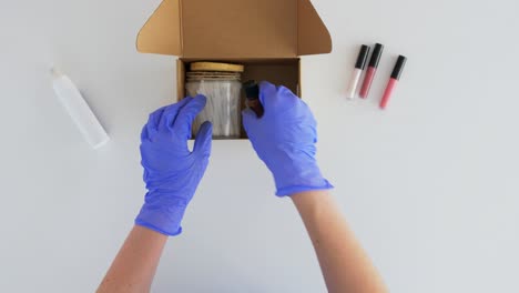 Hands-in-Gloves-Packing-Parcel-Box-with-Cosmetics.delivery,-shipping-and-pandemic-concept--hands-in-protective-medical-gloves-packing-parcel-box-with-cosmetics-and-beauty-products