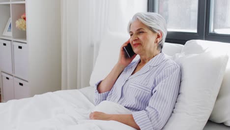 Senior-Woman-Calling-on-Smartphone-in-Bed-at-Home.old-age,-technology-and-people-concept--happy-smiling-senior-woman-in-pajamas-calling-on-smartphone-sitting-in-bed-at-home-bedroom