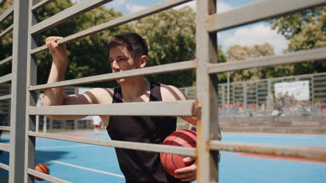 Portrait-of-athletic-serious-man-playing-basketball-posing-alone-stands-behind-grid-on-urban-court