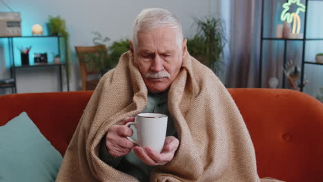 Sick-senior-grandfather-man-wrapped-in-plaid-sit-alone-shivering-from-cold-on-sofa-drinking-hot-tea