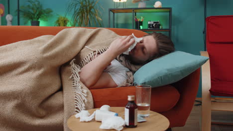 Sick-ill-young-man-suffering-from-cold-or-allergy-lying-on-home-sofa-sneezes-wipes-snot-into-napkin