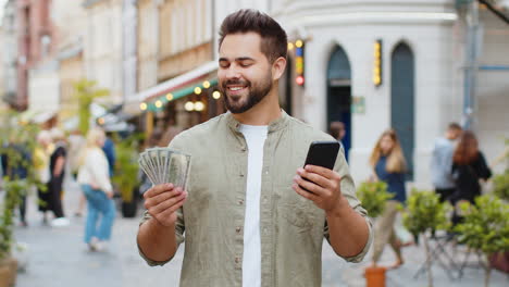 Happy-young-man-counting-money-dollar-cash,-use-smartphone-calculator-app-in-urban-city-street