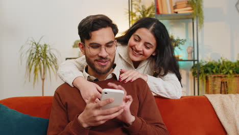 Smiling-Indian-woman-talking-with-boyfriend-swiping-smartphone-sitting-on-sofa-at-home-family-couple