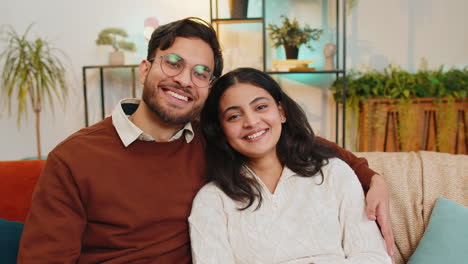 Loving-happy-smiling-young-Indian-couple-spending-leisure-time-sitting-arm-around-relaxing-on-sofa