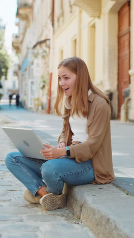 Caucasian-young-woman-using-laptop-computer-celebrating-win-good-message-news-sitting-in-city-street