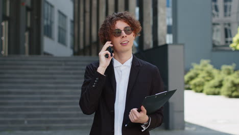 Happy-young-businessman-talking-on-smartphone-on-city-street-near-office-holding-documents-clipboard