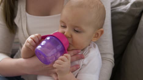 Close-Up-of-Mother-with-Baby-Drinking-Water.family,-motherhood-and-people-concept-close-up-of-mother-with-little-baby-drinking-water-from-sippy-cup-at-home